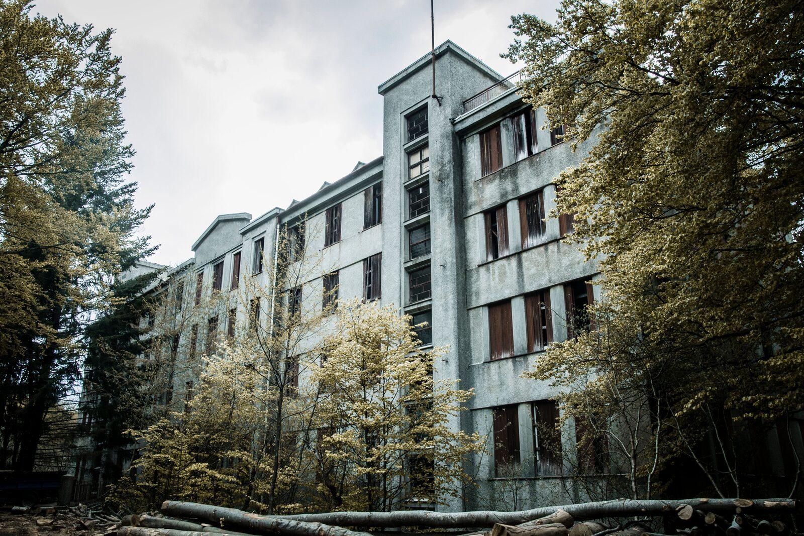 An abandoned four-storey hospital building, windows broken and dark, framed by overgrown trees